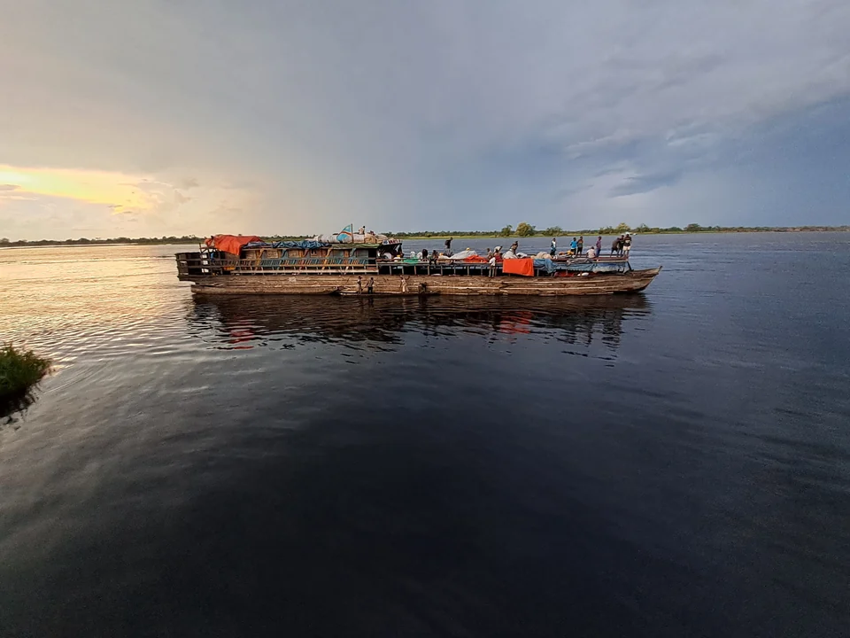 A loaded wooden cargo boat crossing the calm, wide Congo River at Mbandaka under a stormy sky