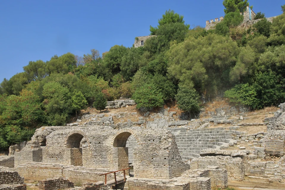 Ruined stone arches and tiered seats of the ancient theater at Butrint, Albania, below a wooded hill with a castle