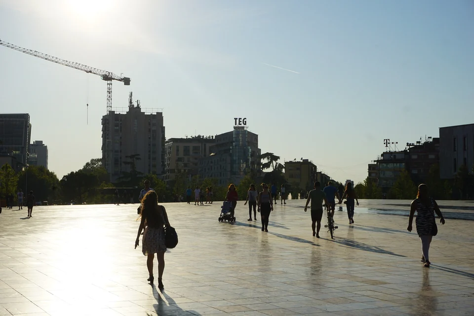 People walking across the wide stone paving of Skanderbeg Square in Tirana in late afternoon sun