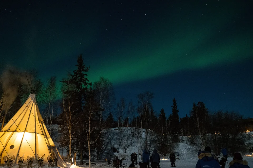 Green aurora borealis over snowy boreal forest and a glowing tipi near Yellowknife, Northwest Territories
