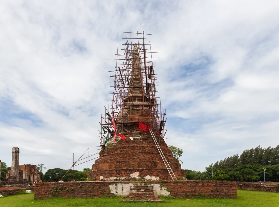 Backstein-Stupa im Wat Mahathat in Ayutthaya, in Gerüste gehüllt, dahinter Ruinenmauern und Bäume
