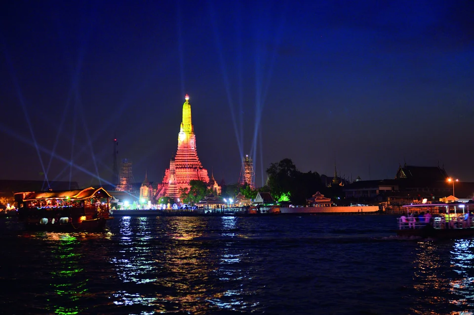 Der beleuchtete zentrale Prang des Wat Arun am Chao Phraya bei Nacht, mit einem vorbeifahrenden Touristenboot