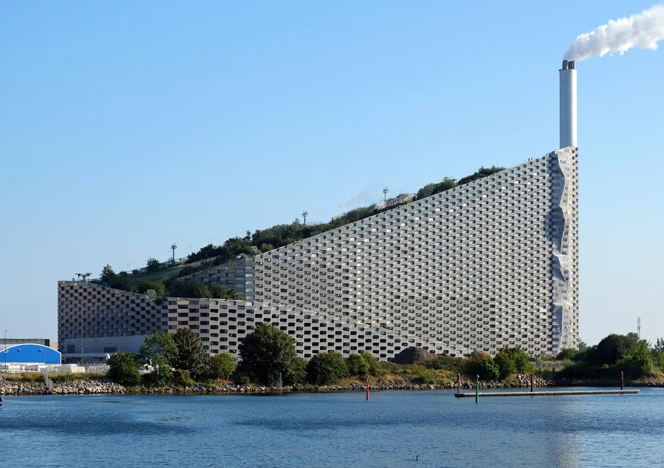Copenhill power plant in Copenhagen with a green rooftop ski slope, slanted facade and chimney beside the water
