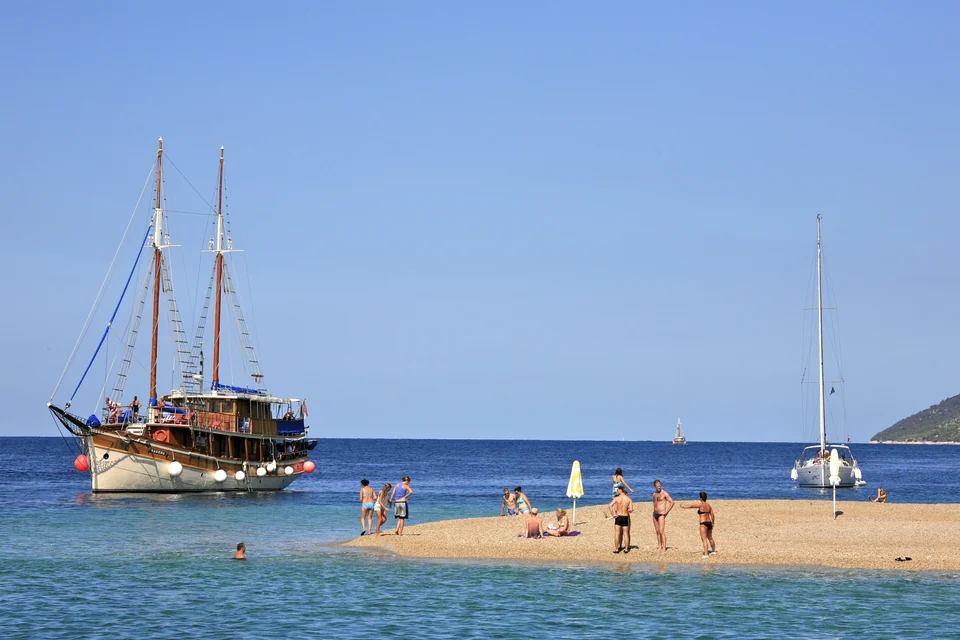 Swimmers on the pebble tip of Zlatni Rat beach near Bol on Brač, with a wooden sailing ship anchored offshore