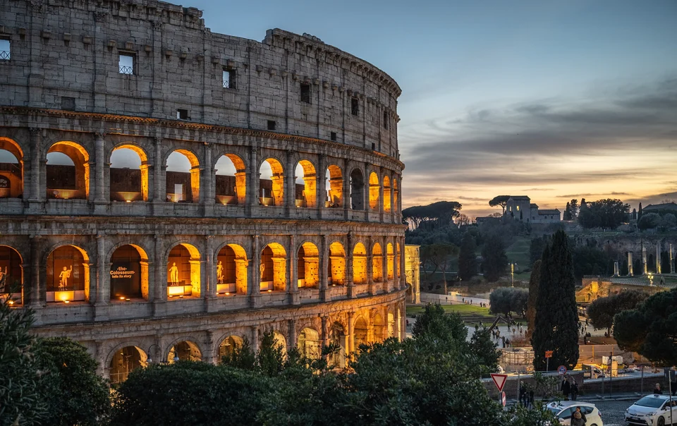 The illuminated arches of the Colosseum in Rome at dusk with trees and the evening sky