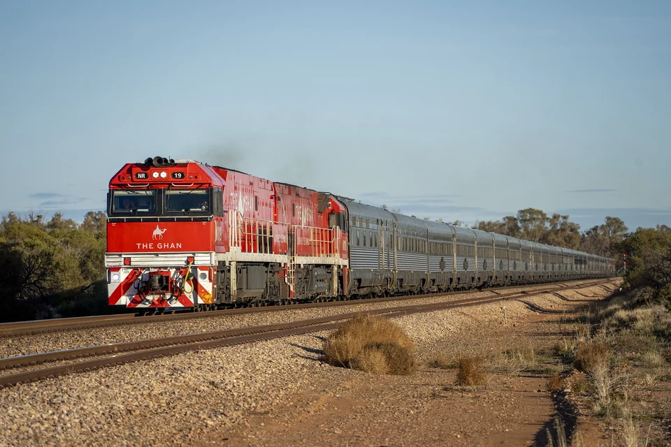 Red locomotive pulling the long silver Ghan passenger train through dry scrubland in South Australia