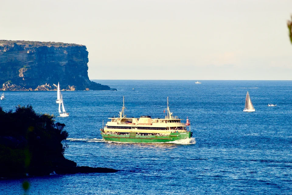 Green and yellow Manly ferry crossing Sydney Harbour near North Head with sailboats in the background