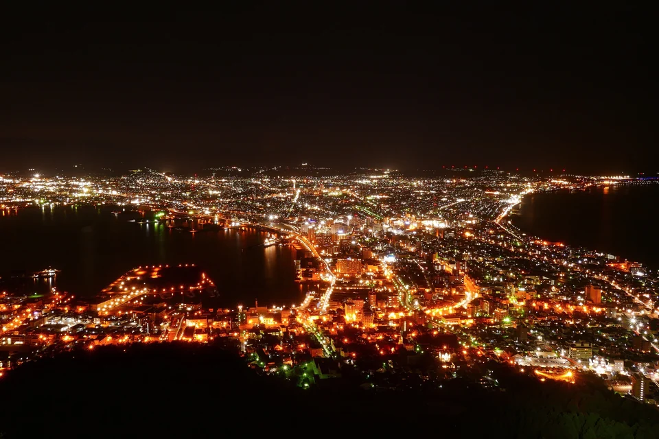 Die erleuchtete Stadt Hakodate und ihre geschwungene Küstenlinie bei Nacht, gesehen vom Gipfel des Hakodate-Bergs
