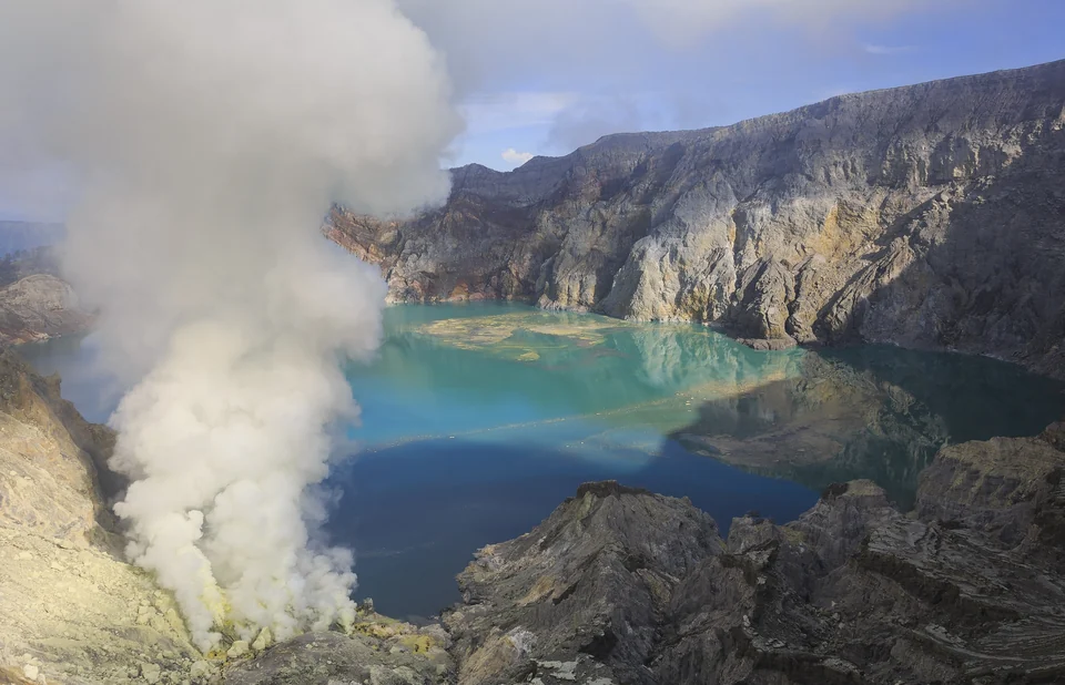 Turquoise acidic lake on the floor of the Kawah Ijen crater with steep gray crater walls and drifting volcanic gas