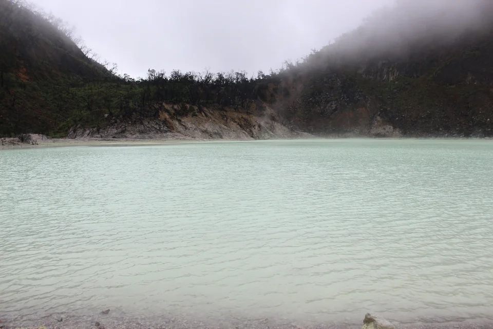 Milky pale green acidic water of Kawah Putih crater lake surrounded by white rocks and forested slopes near Bandung