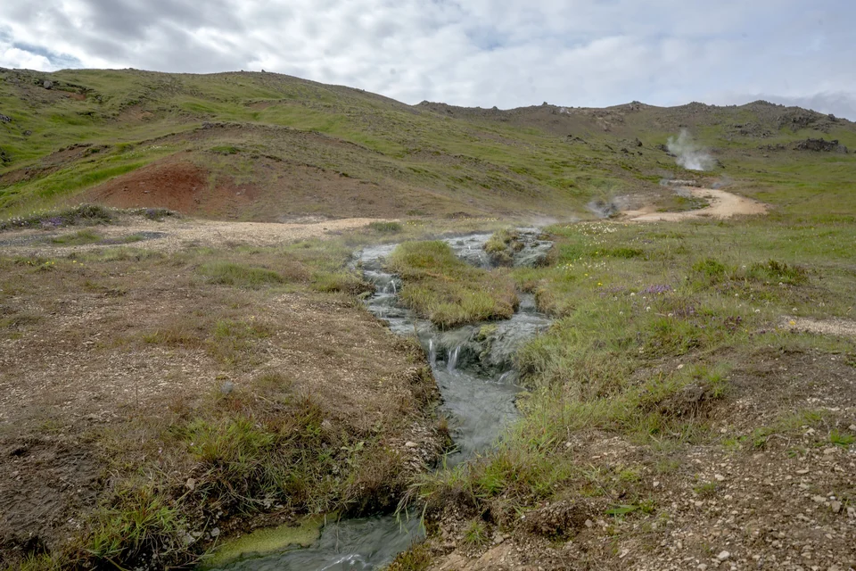 Warm geothermal stream flowing through green hills with steam vents rising behind in Reykjadalur valley near Hveragerði