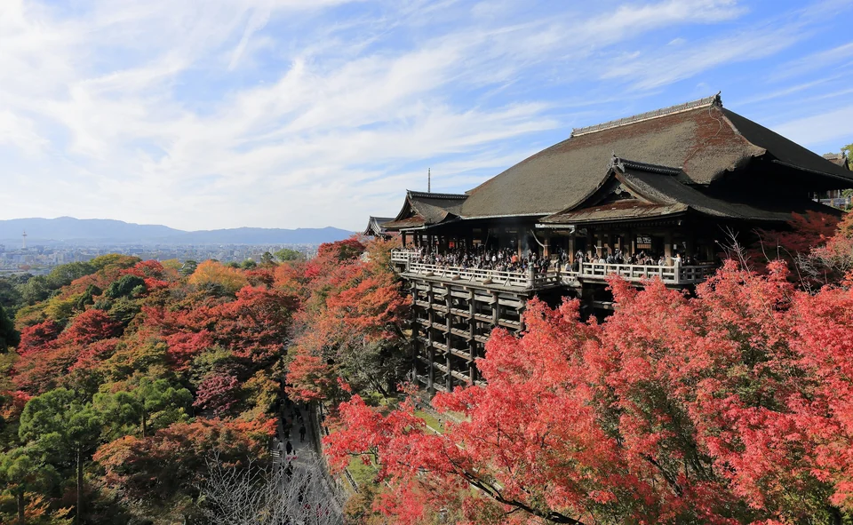 Kiyomizu-dera temple’s wooden stage above red autumn maples, with the Kyoto skyline and mountains behind