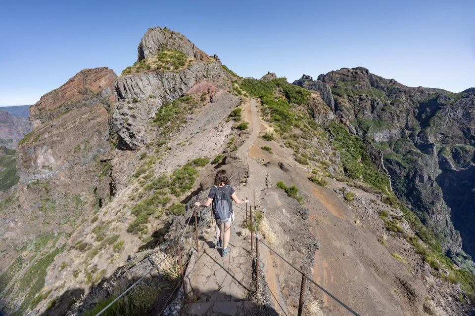 A hiker descends a stone path with railings along the PR1 ridge between Pico do Arieiro and Pico Ruivo, Madeira