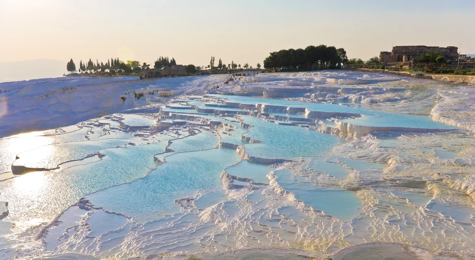 Turquoise travertine pools on the terraces of Pamukkale at sunset, with trees and old stone walls on the ridge beyond
