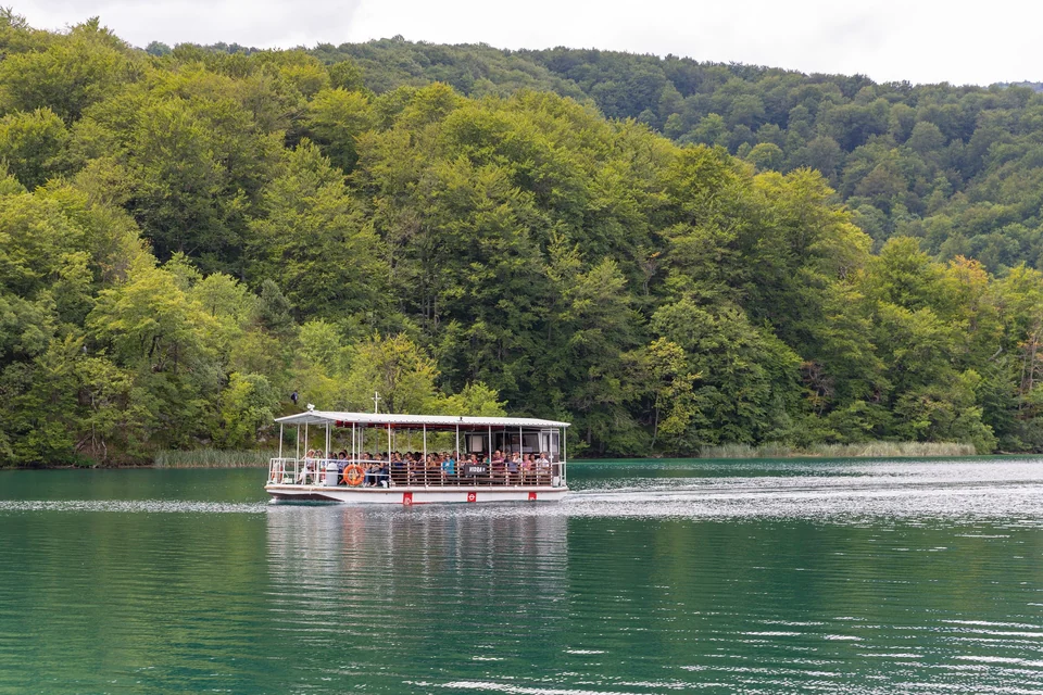 Barco eléctrico de pasajeros abierto cruzando el lago Kozjak bajo colinas boscosas en el Parque Nacional de los Lagos de Plitvice