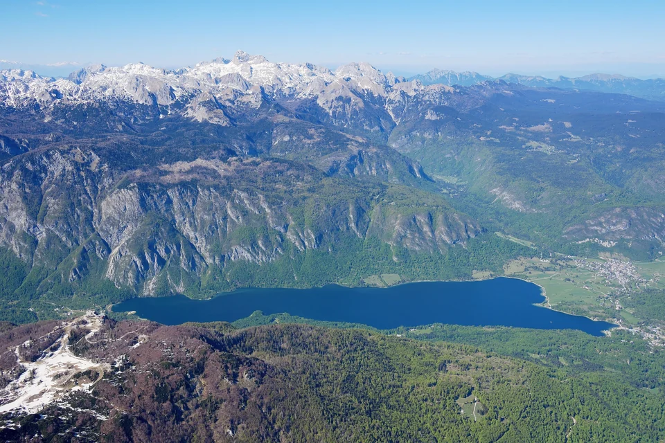 Aerial view of Lake Bohinj surrounded by forested mountains, with Mount Triglav in the distance
