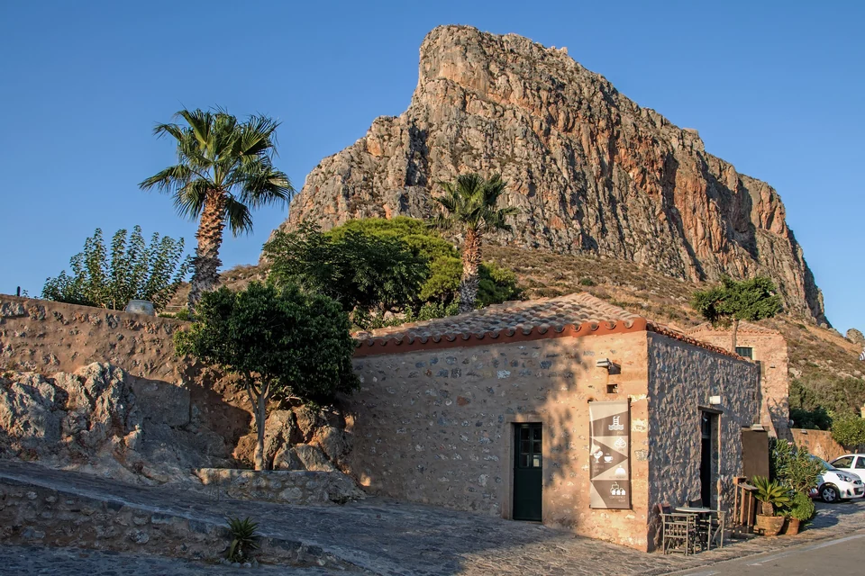 The massive rock of Monemvasia towering behind a stone house and palm trees on the Peloponnese coast