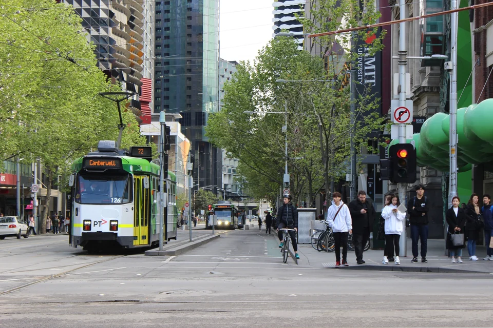 Green and white tram on tree-lined Swanston Street in Melbourne with pedestrians and a cyclist