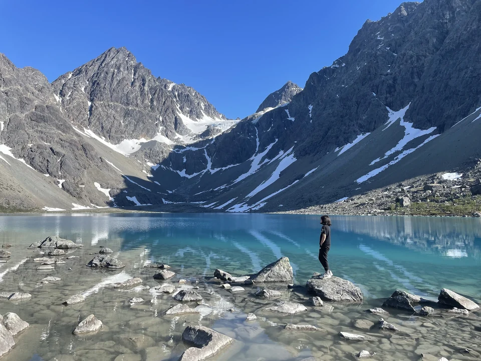Hiker standing on a rock at the edge of turquoise Blåisvatnet lake below snowy peaks in the Lyngen Alps, Norway